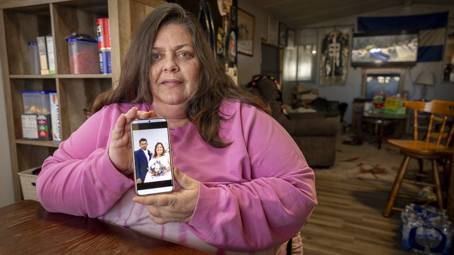 Stacey Murillo Martinez holds a smart phone with a vertical wedding portrait of her and husband Miguel on its screen while sitting at a wood table in a room with wood flooring and with shelves with food containers in the background on one side and another room with chairs, lamps, wall art and other items to the other side.
