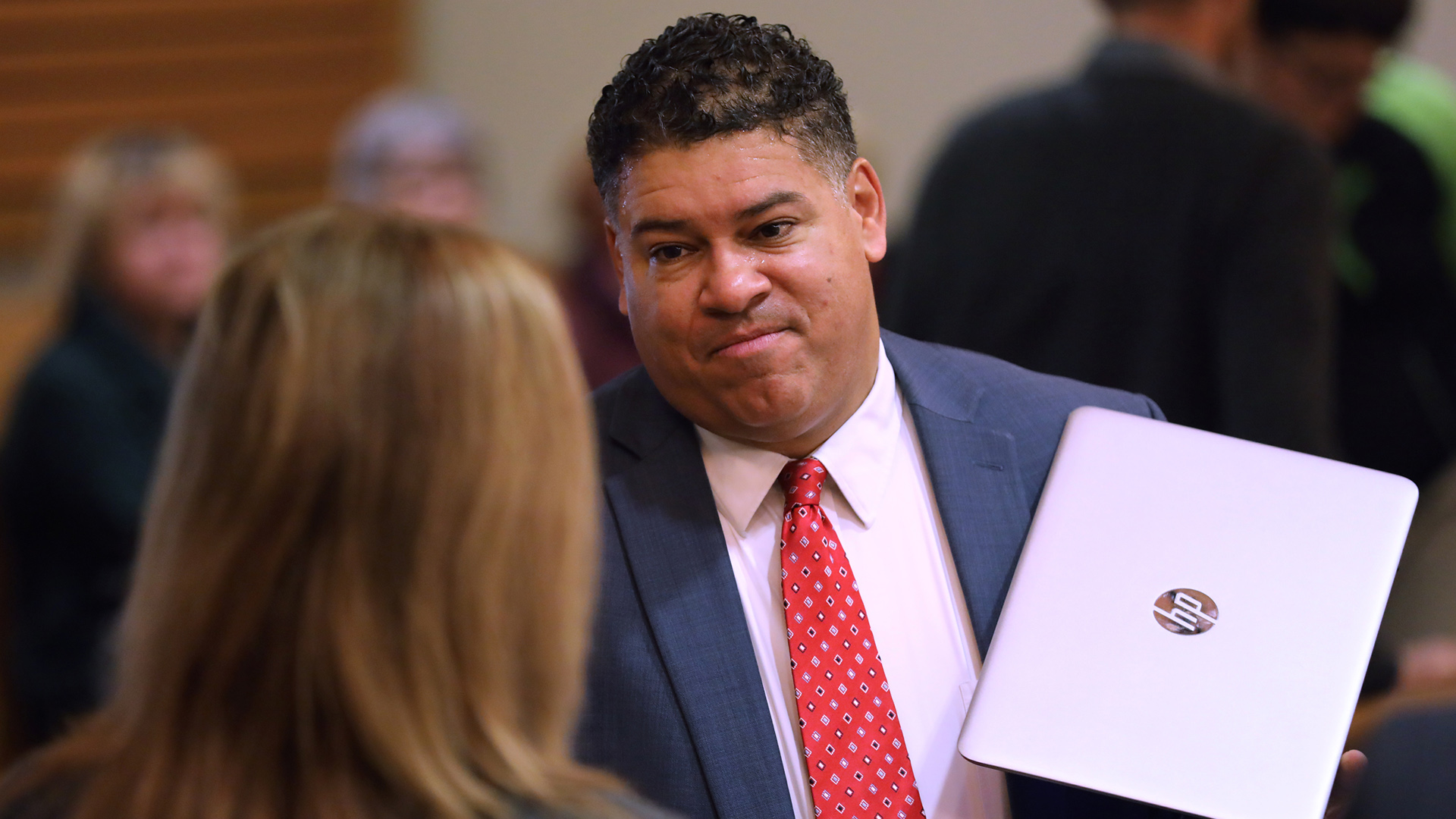 Ismael Ozanne stands and holds a laptop computer next to his chest with his left hand while speaking with an out-of-focus person in the foreground, with several other out-of-focus people seated and standing in the background.
