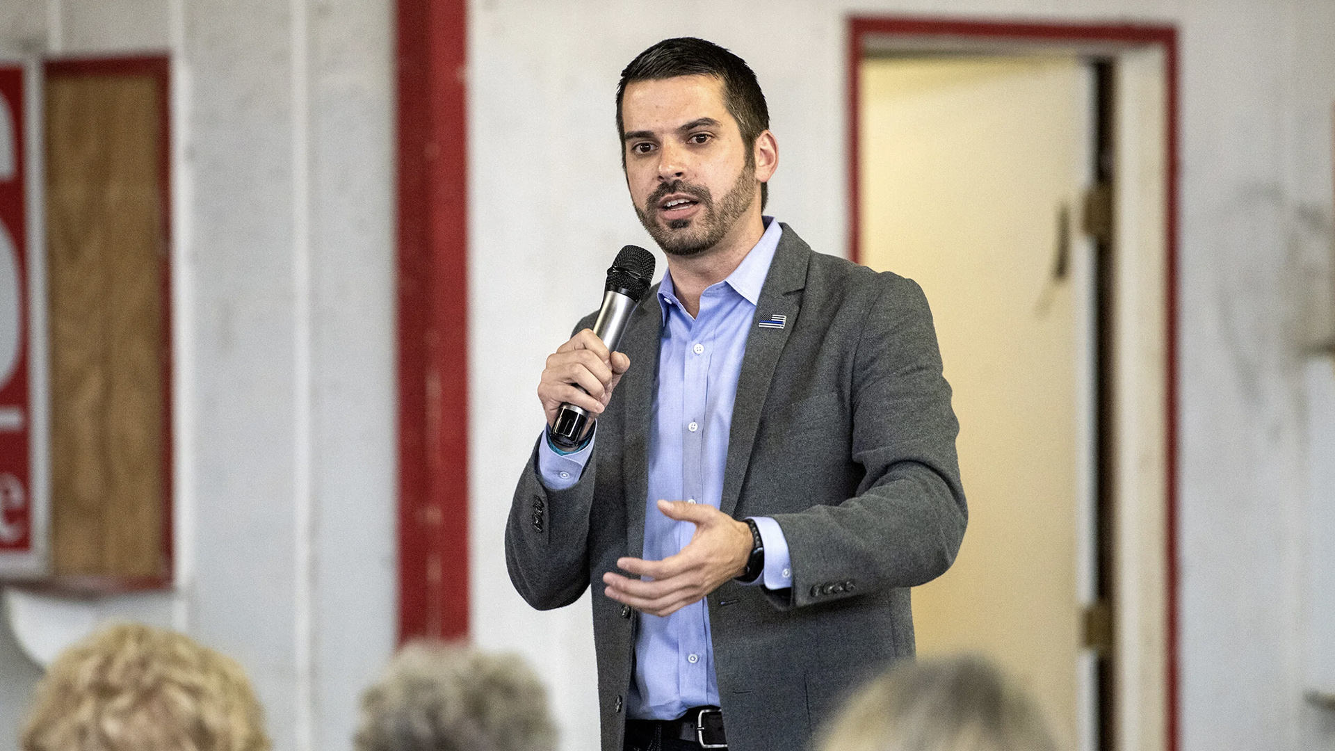 Eric Toney speaks and holds a wireless microphone in his right hand and gestures with this left hand while stand in a room with an open door in the background and the out-of-focus heads of three people seated in the foreground.