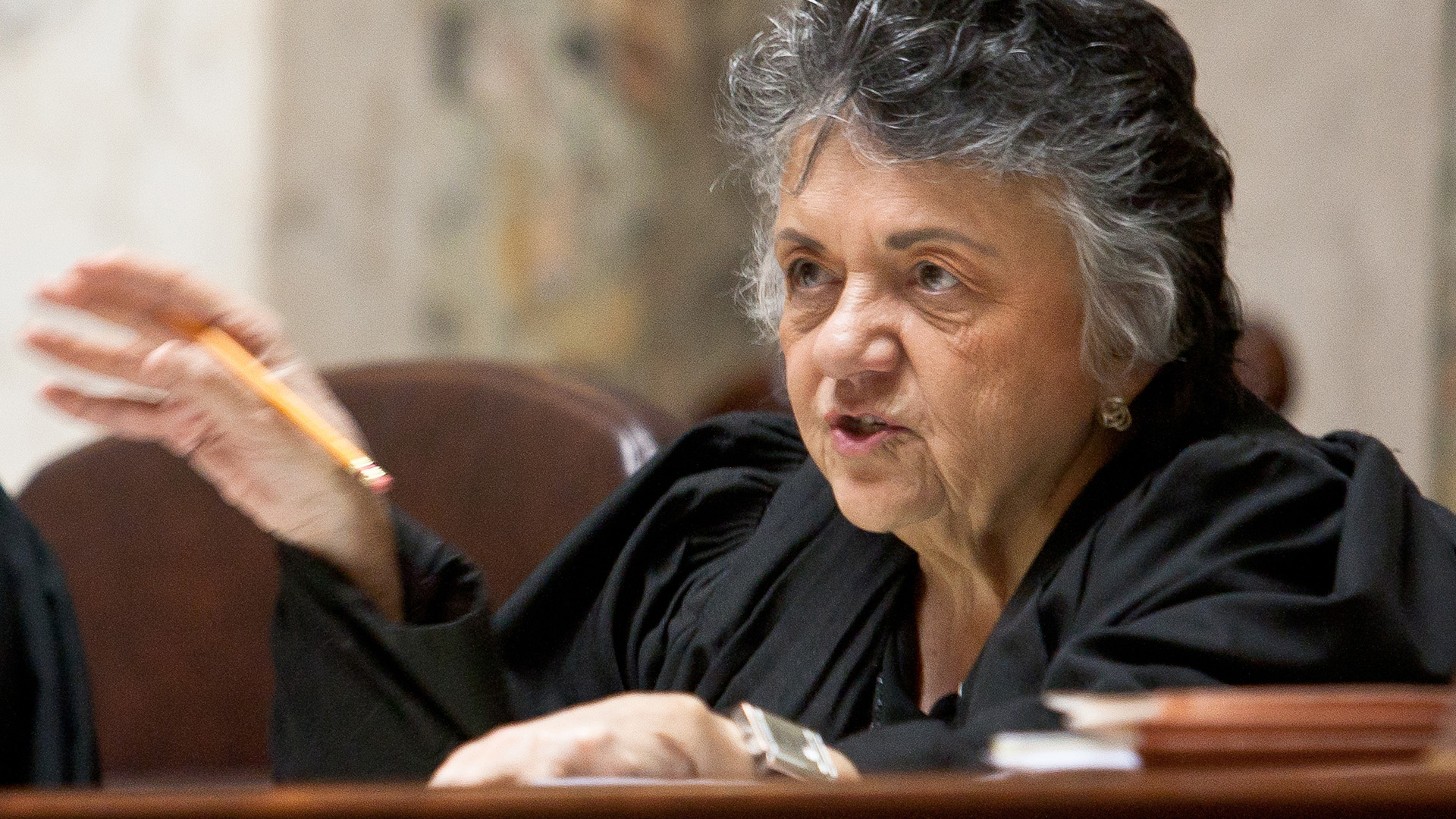 Shirley Abrahamson raises her right hand in-motion while holding a pencil between her thumb and forefinger, while sitting a judicial bench with her out-of-focus left hand and multiple books on its surface.