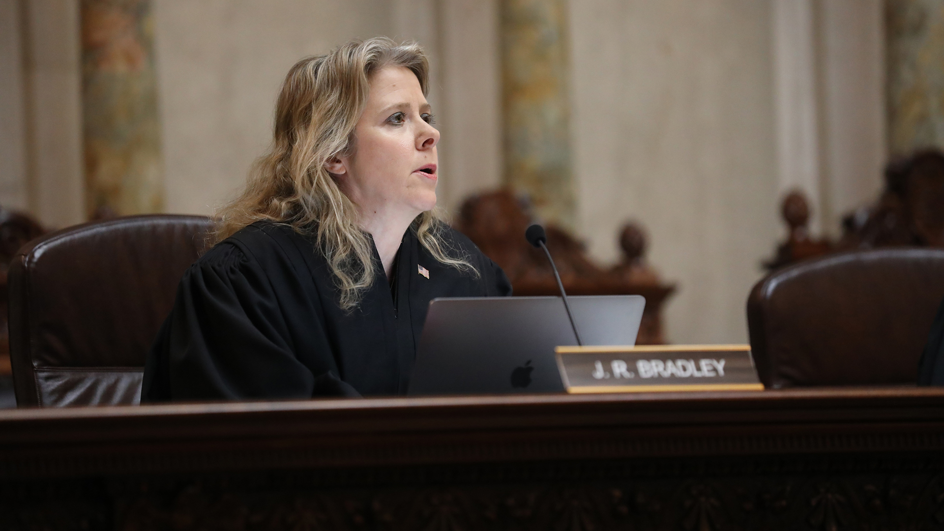 Rebecca Bradley listens while sitting in a high-backed leather chair behind a judicial dais with a name plate on its surface reading "J.R. Bradley," with out-of-focus high-backed wood chairs and marble masonry in the background.
