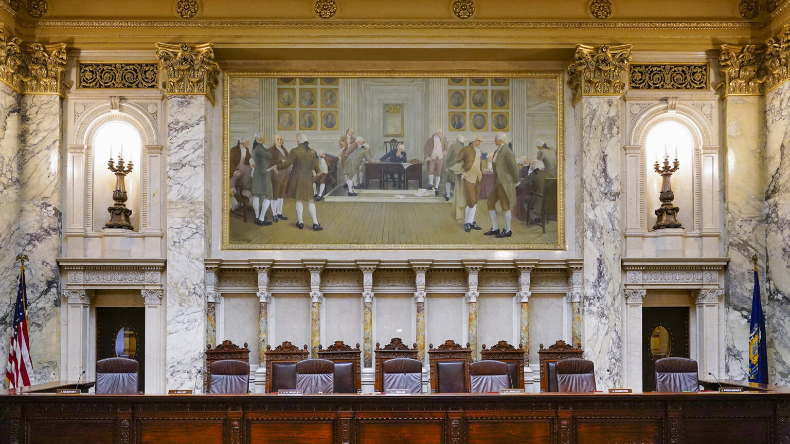 A judicial bench with two sets of seats, one leather and another wood and leather, is between the U.S. and Wisconsin flags and below a large painting mounted on a wall with marble masonry and illuminated brass light fixtures in wall niches. A judicial bench with two sets of seats, one leather and another wood and leather, is between the U.S. and Wisconsin flags and below a large painting mounted on a wall with marble masonry and illuminated brass light fixtures in wall niches.