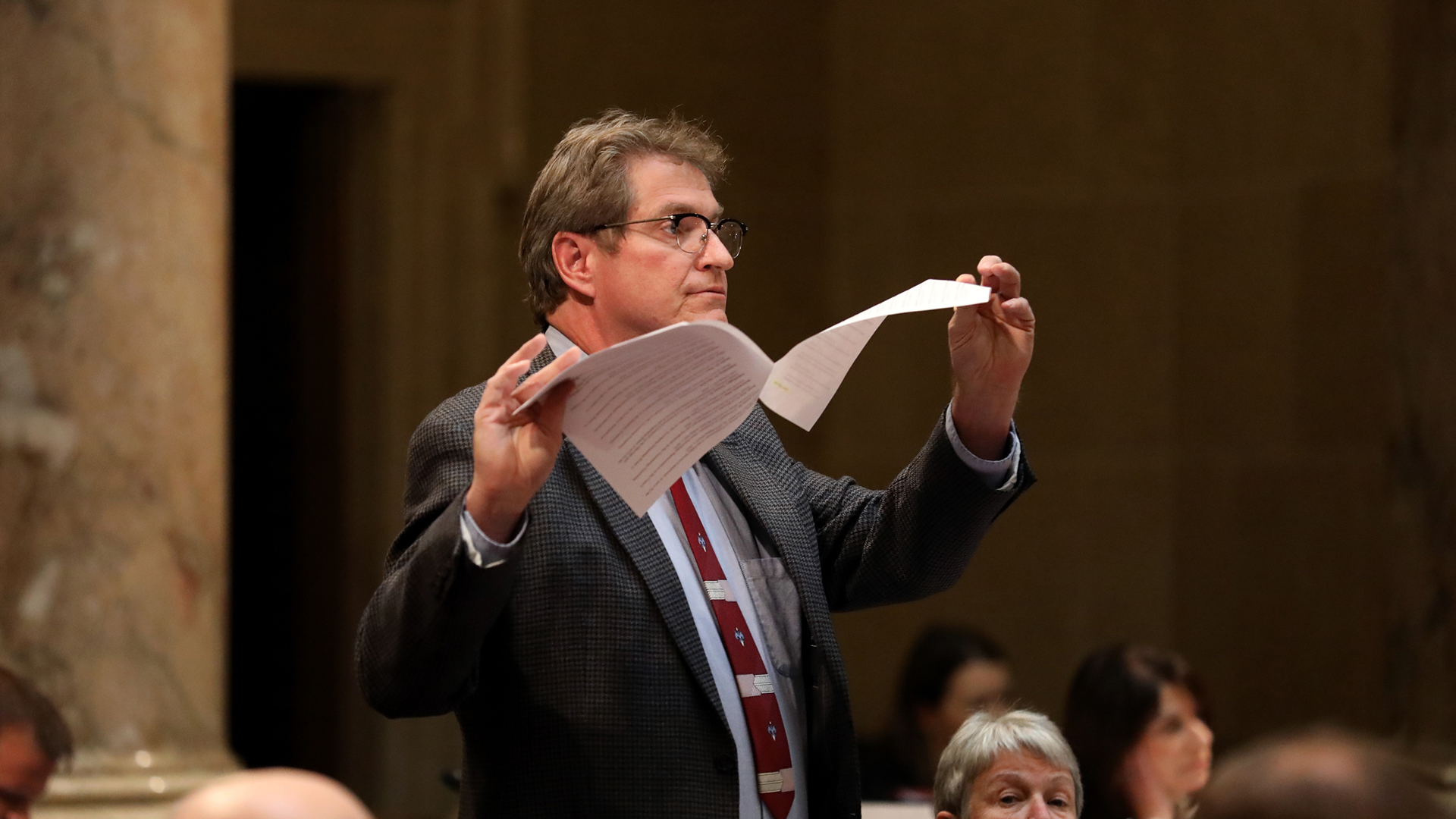 Tim Carpenter holds pieces of paper connected by a staple in both hands while standing in a room with people seated around him and out-of-focus marble masonry and a pillar in the background.