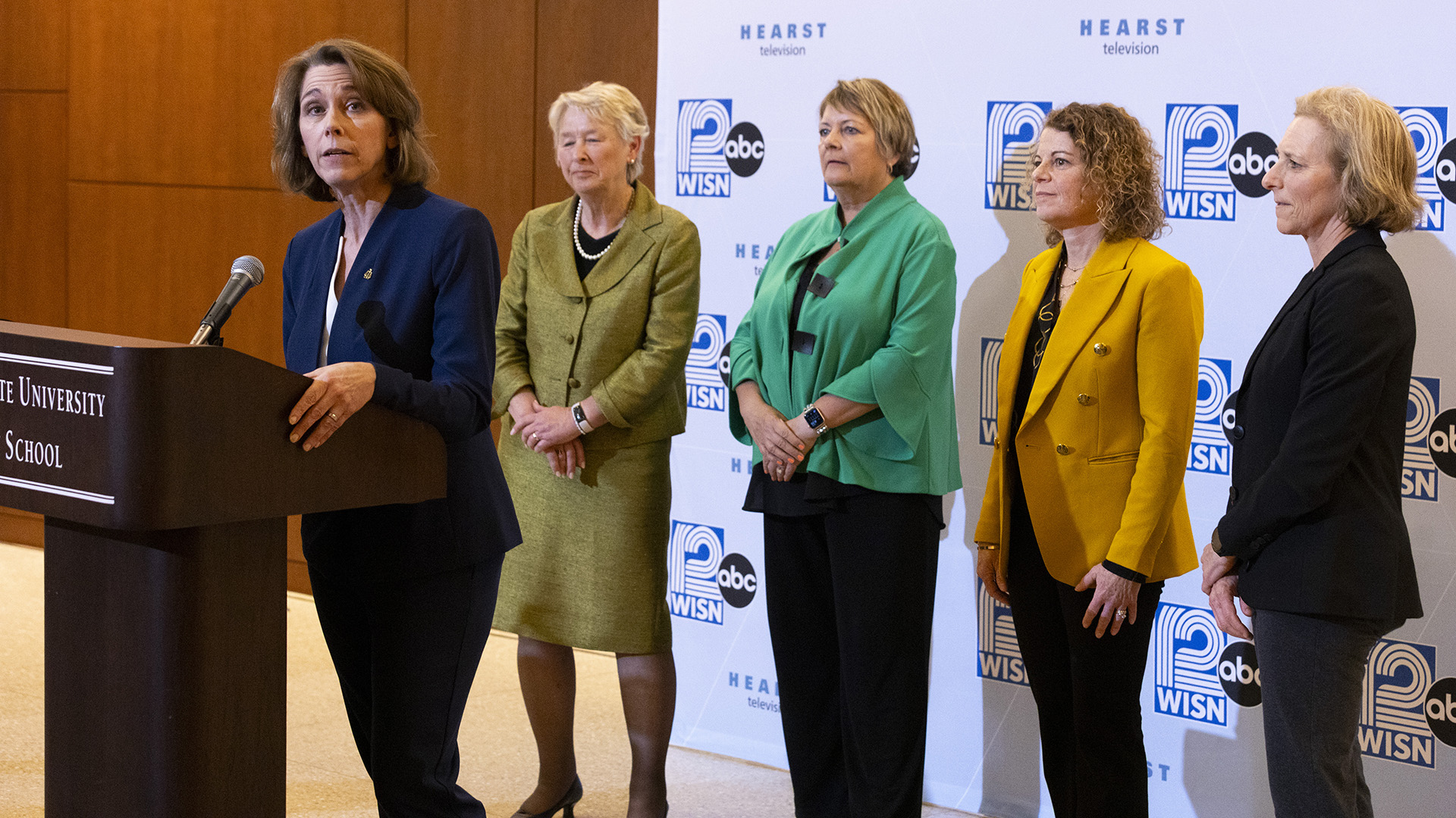 Susan Crawford speaks while standing in front of a wood podium with a microphone mounted on its surface, with Ann Walsh Bradley, Janet Protasiewicz, Rebecca Dallet and Jill Karofsky standing behind her in front of a logo backdrop, in a room with wood-paneled walls.