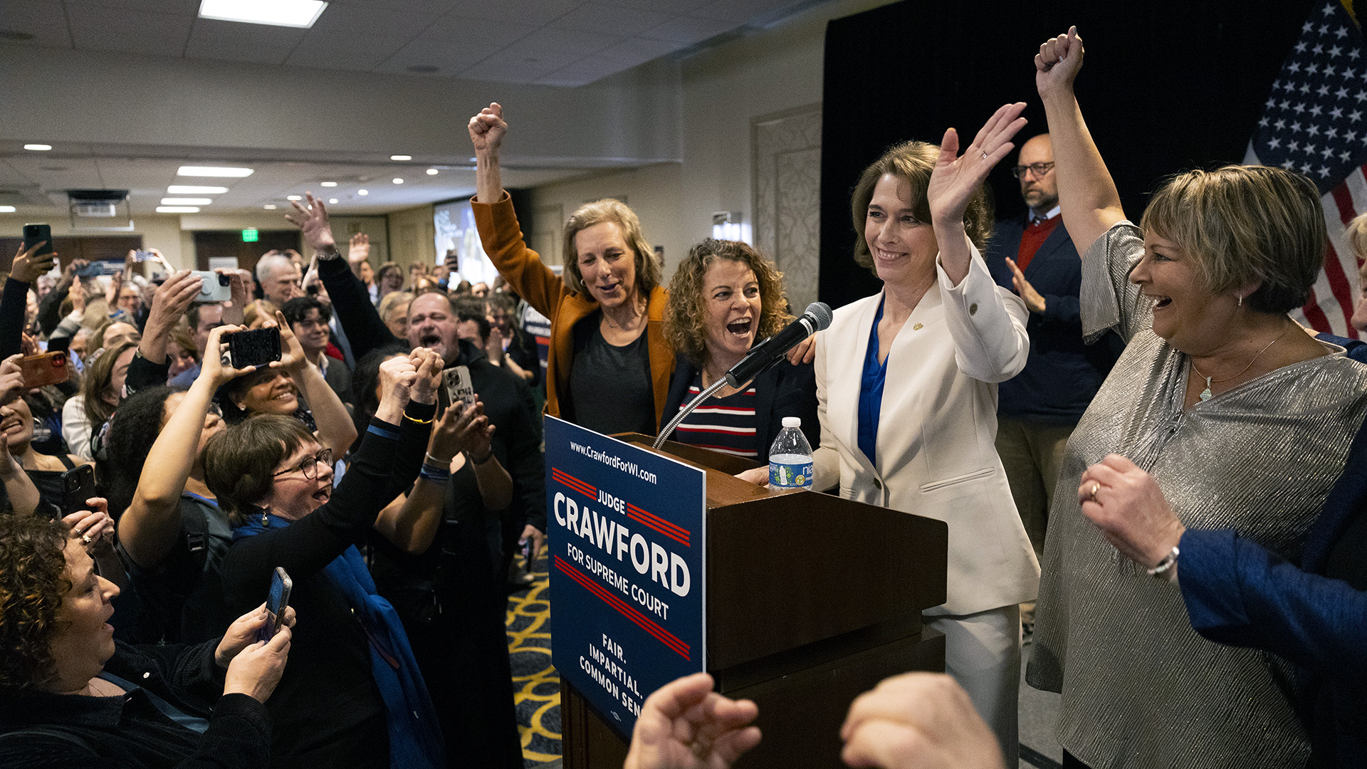 Susan Crawford waves with her left hand while standing behind a wood podium with a microphone mounted to its top and a campaign sign affixed to its front, with Jill Karofsky and Rebecca Dallet standing to one side and Janet Protasiewicz standing to the other side in front of a pipe-and-drape backdrop and a U.S. flag, with cheering people standing in front of them, with many holding up phones to record video and take photos, in a room with fluorescent lighting in a multi-level ceiling.