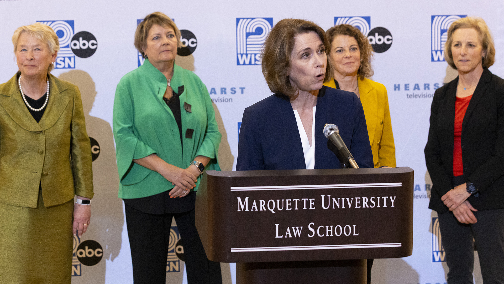 Susan Crawford speaks while standing in front of a wood podium with a microphone mounted on its surface, with Ann Walsh Bradley, Janet Protasiewicz, Rebecca Dallet and Jill Karofsky standing behind her in front of a logo backdrop.