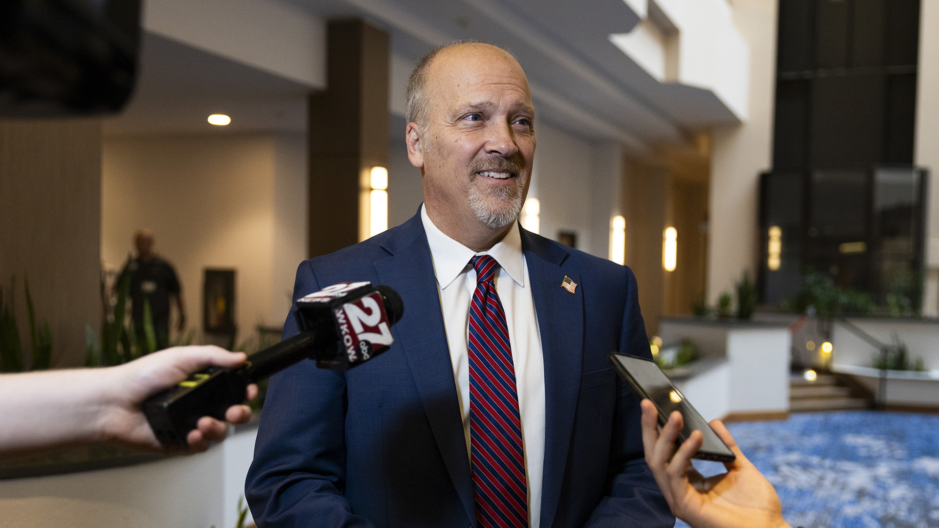 Brad Schimel speaks to two people holding a wireless microphone with the flag of a media organization and a smart phone in front of him, while standing in an atrium with a hallway to one side and a sunken level on another side.