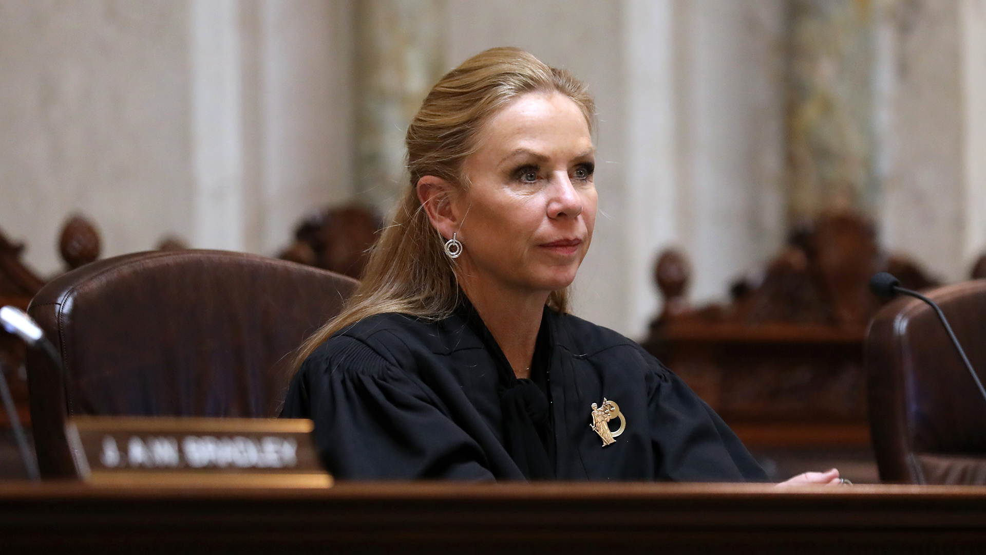 Annette Ziegler listens while sitting in a high-backed leather chair behind a judicial dais, with other high-backed wood chairs in the background.