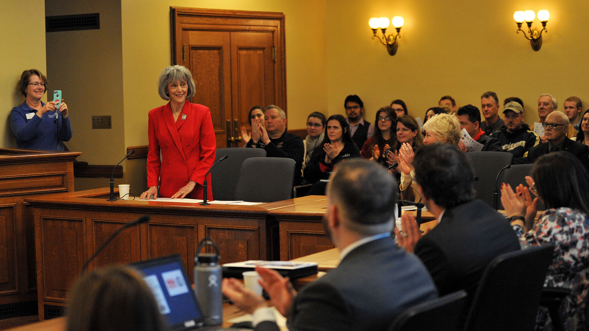 Eve Galanter stands in front of chairs in a wood witness desk with other people seated at tables in the foreground and in a gallery in the background applauding her, with a person standing next to a wall recording the scene with a smart phone, in a room with wood double doors and illuminated brass wall sconce light fixtures in the background.
