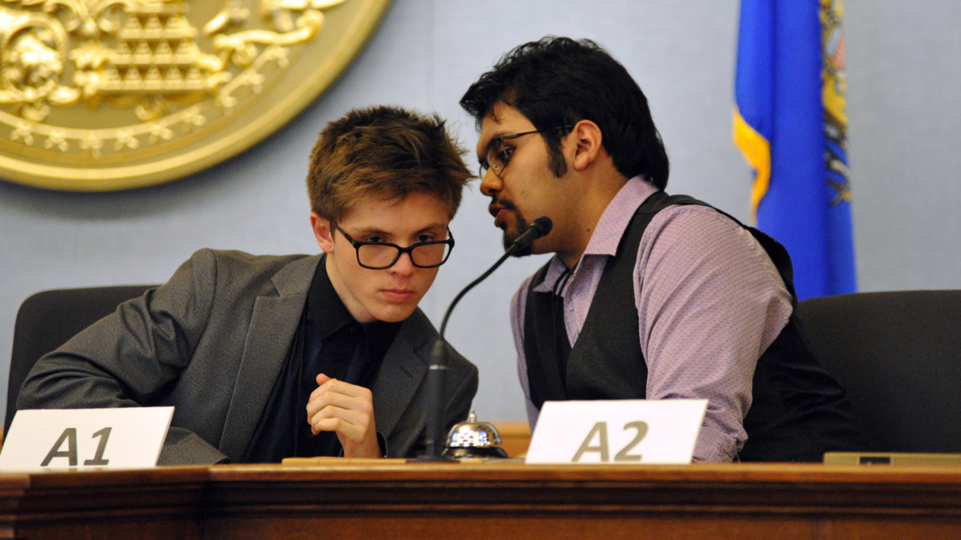 Seth Mayrer and Carlos Herrade lean toward each other and speak while seated at a wood legislative dais with a microphone affixed to its surface alongside a table bell and two paper signs that read "A1" and "A2," in a room with a wall-mounted Great Seal of the State of Wisconsin and a Wisconsin flag in the background.