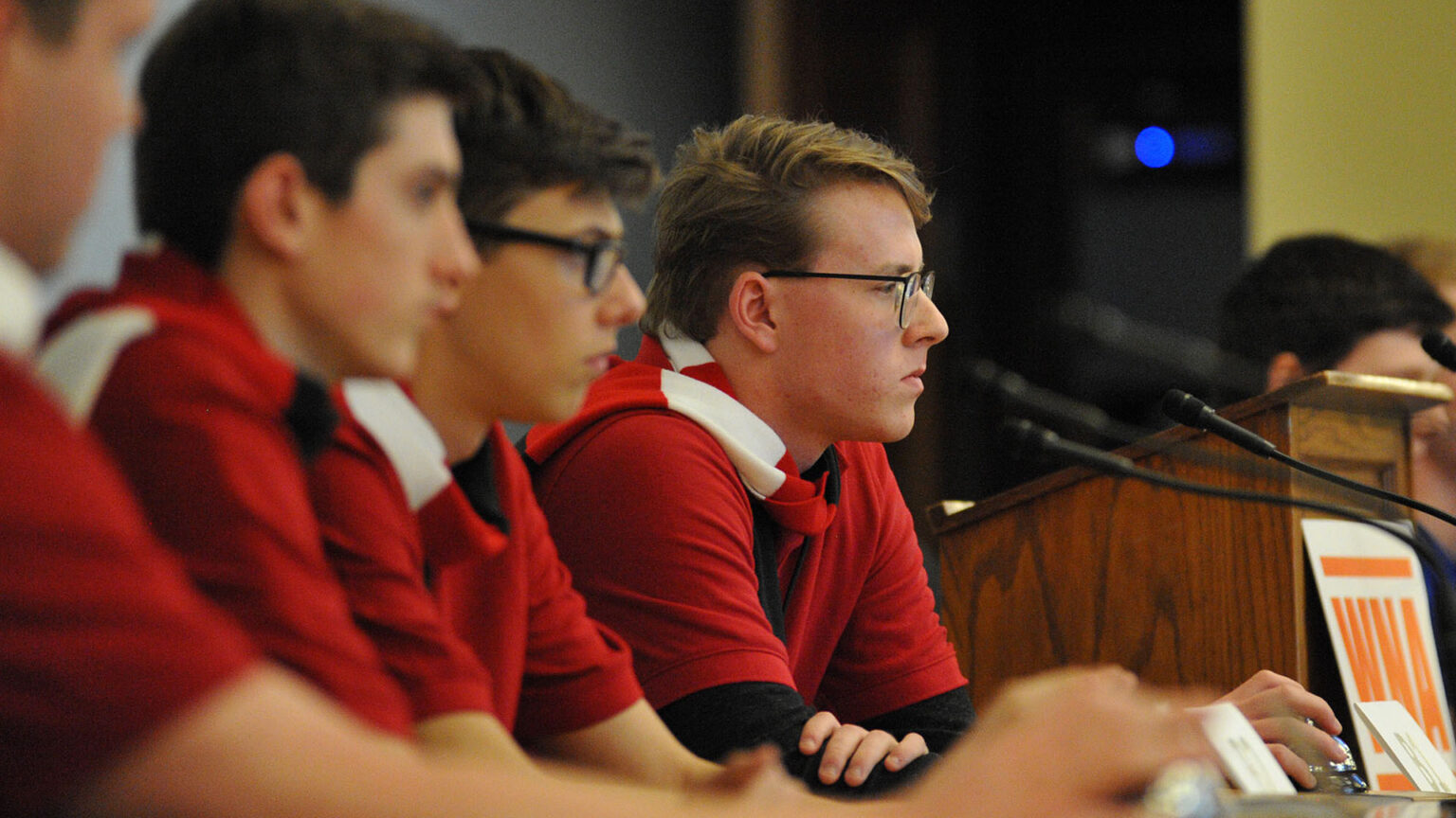 Kevin LeGrow and three out-of-focus people seated to his right sit at a table facing mounted microphones, with a wood podium and other people seated on its far side in the background.