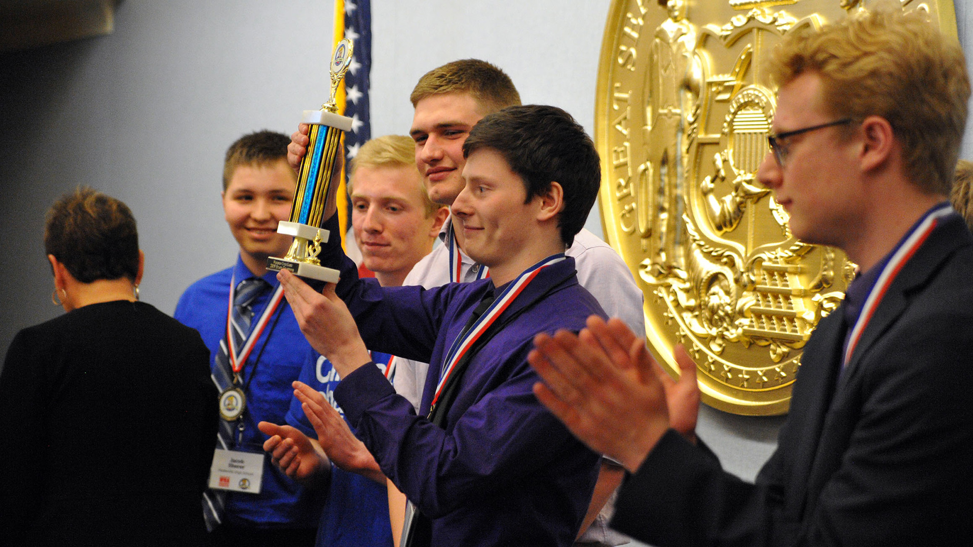 Liam Reinicke holds a trophy in the air with both hands whild standing between other people who are applauding, in a room with a U.S. flag and a wall-mounted Great Seal of the State of Wisconsin in the background.