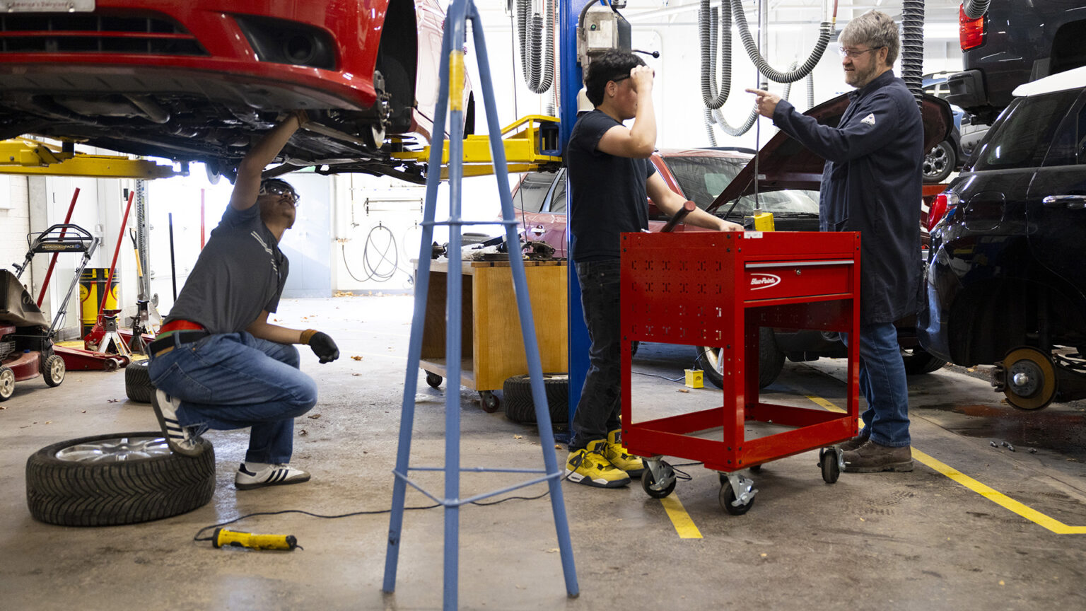 Miles Tokheim points his left index figure while facing José Ruiz as both stand on either side of a rolling tool chest, with Oscar Haro Rodriguéz crouching with this right foot on a tire resting on its side while under the bottom of a vehicle with detached tires that is elevated on a car lift, in a room with a concrete floor and other vehicles and equipment in the background.