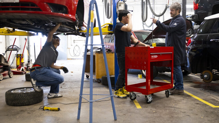 Miles Tokheim points his left index figure while facing José Ruiz as both stand on either side of a rolling tool chest, with Oscar Haro Rodriguéz crouching with this right foot on a tire resting on its side while under the bottom of a vehicle with detached tires that is elevated on a car lift, in a room with a concrete floor and other vehicles and equipment in the background.