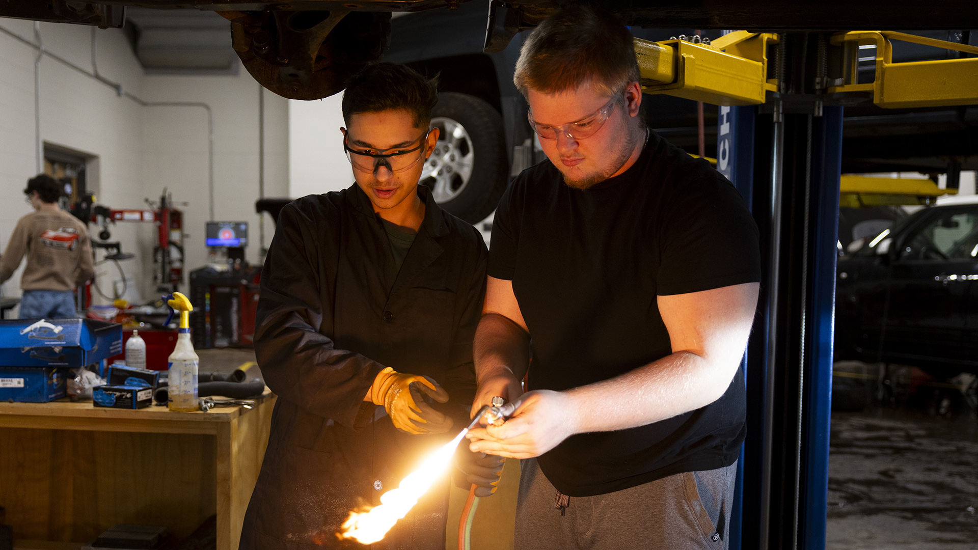 Lucas Pearl holds the handle of an ignited torch with two hands while standing next to Edduar Beperec, who holds its fuel hose with a gloved left hand and reaching toward it with a gloved right hand, with both standing under an elevated vehicle on a car lift, with a wood table with tools on its surface along with other vehicles, equipment and a standing person in the background of a room with a high ceiling.