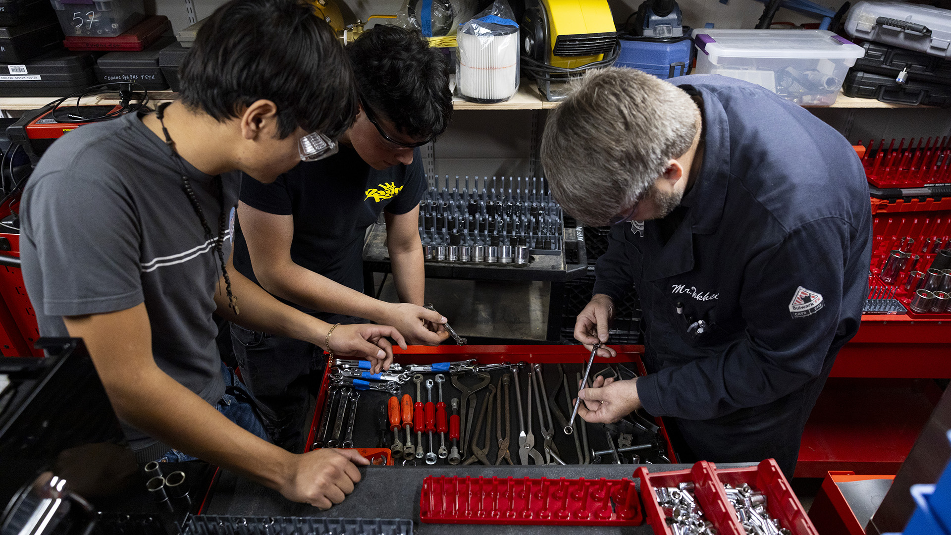 Oscar Haro Rodriguéz, José Ruiz and Miles Tokheim stand and lean over a drawer in a tool cabinet, with other auto part and tool containers on a table in the foreground and on shelves in the background.