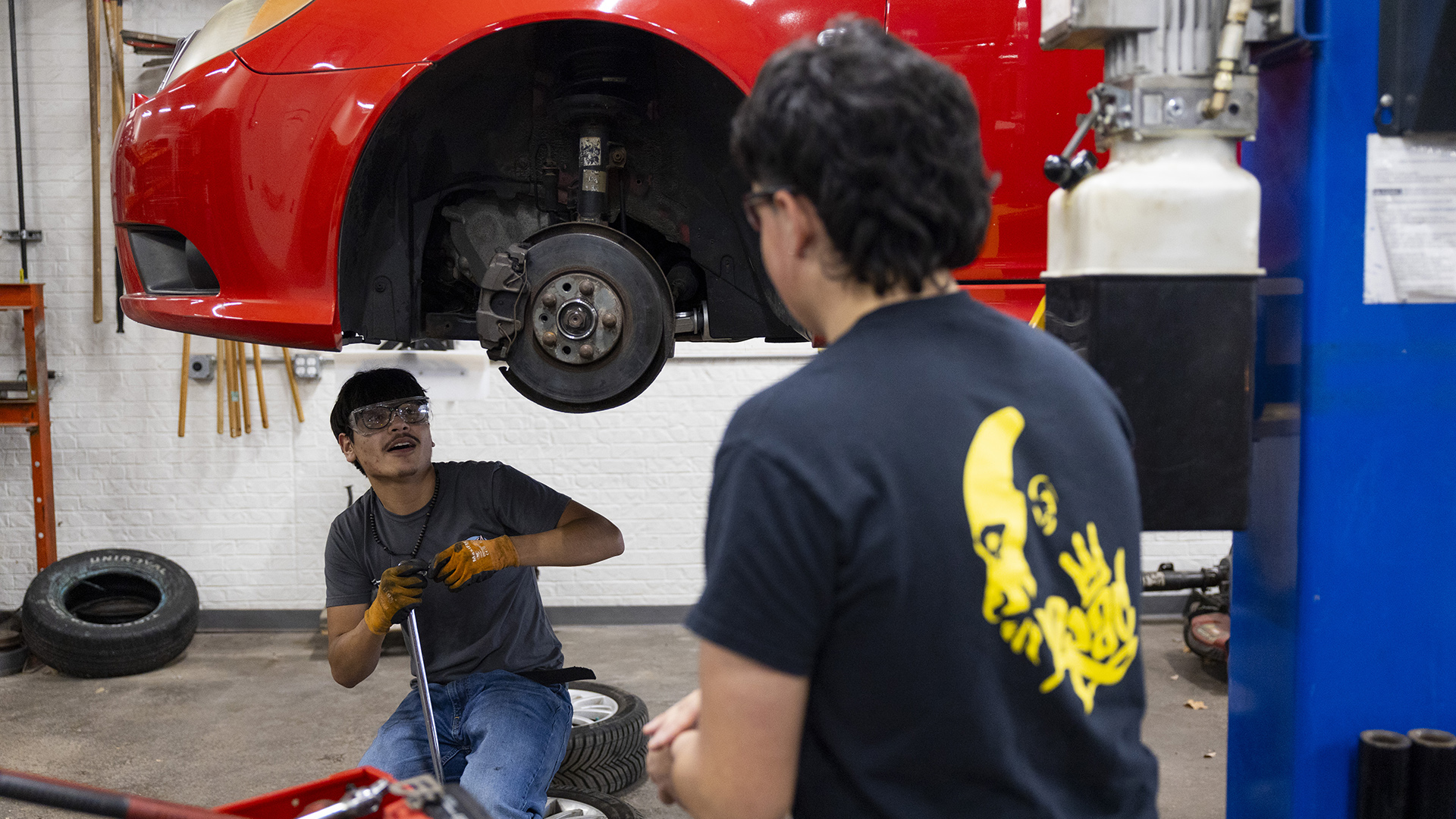 Oscar Haro Rodriguéz holds a tool in his right hand while crouching under a vehicle with no tires that is elevated on a car lift, with multiple tires on a concrete floor and a painted brick wall in the background.