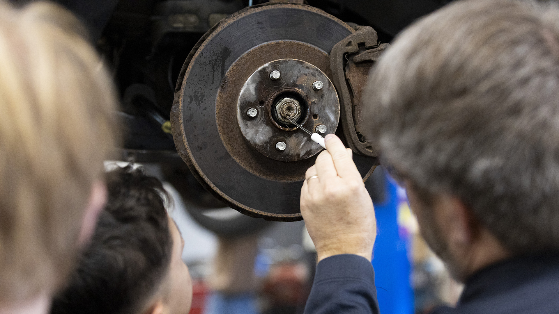 Miles Tokhiem holds a small tool in his left hand and points toward the center of a wheel hub on an elevated vehicle, with the back of his out-of-focus head and two students in the foreground.