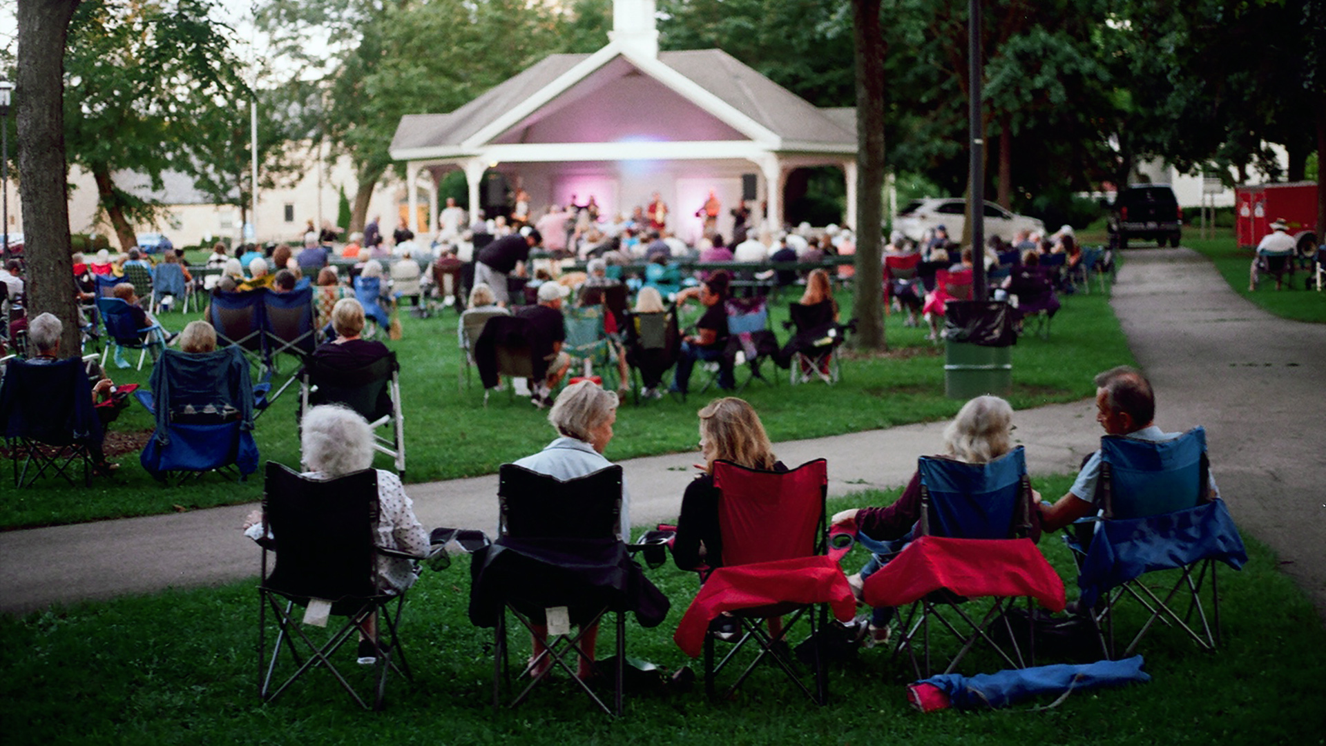 Five people sit in folding camp chairs on a flatiron section of lawn next to a sidewalk intersection and face an out-of-focus open-walled building in the background with musicians performing on a stage, with dozens of other people seated on another section of lawn in front of the structure.