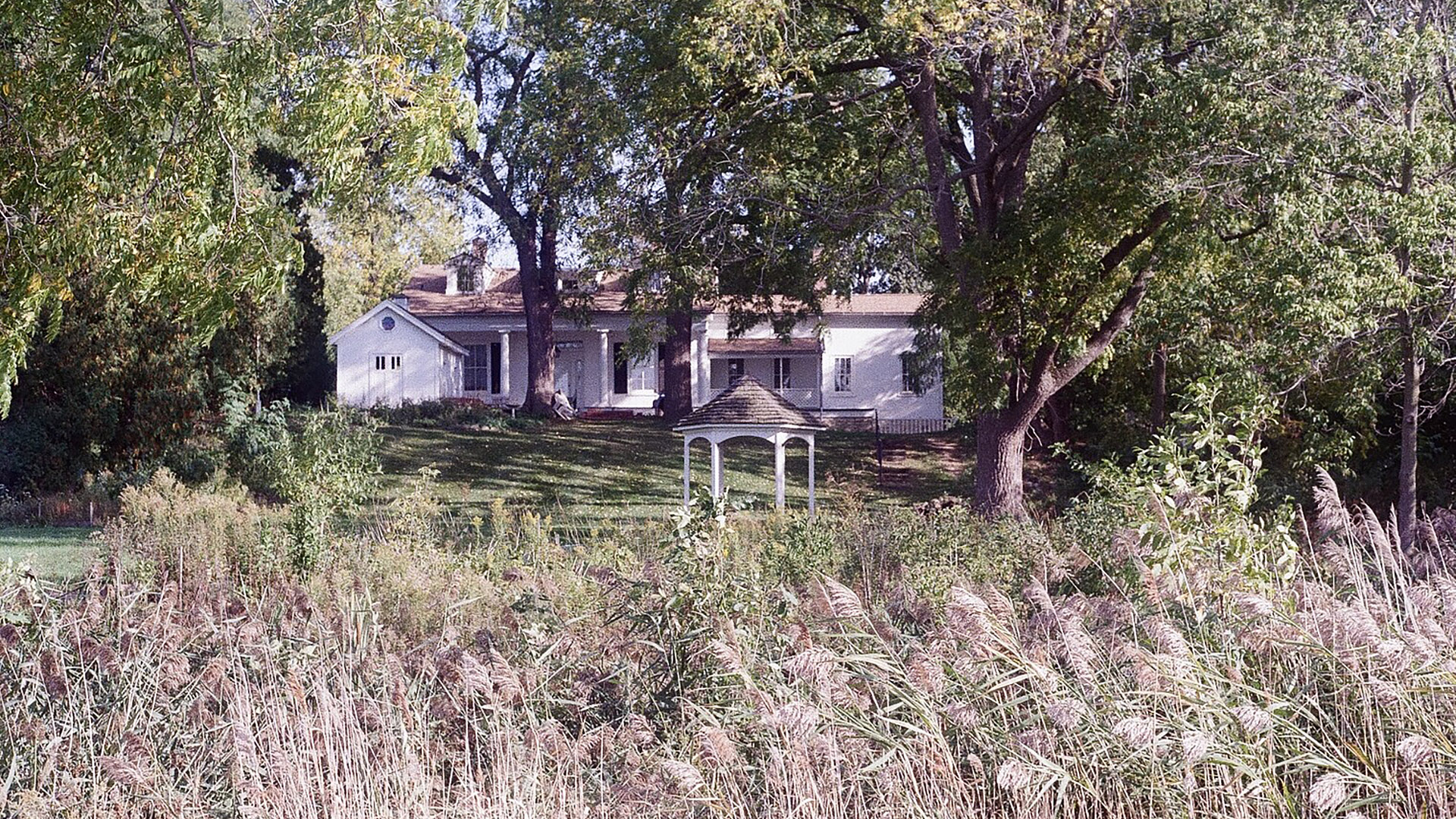 A multi-story house stands among multiple mature trees, with a gazebo standing on a lawn between the building and a stand of prairie plants in the foreground.