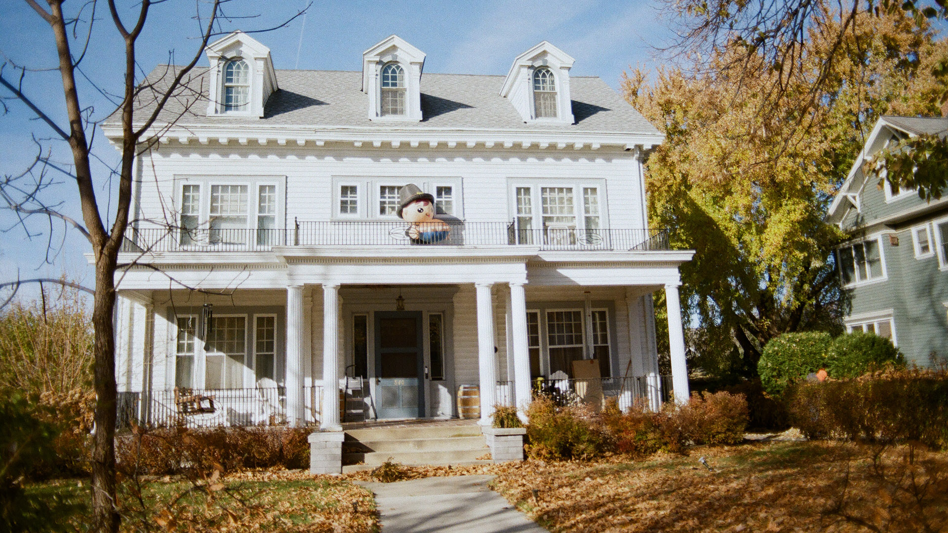 An inflatable figure sits on the front balcony of a three-story house with a colonnaded porch and three dormers on its roof, with fallen leaves covering a front lawn, another house to the side, and trees with different amount of leaves in the foreground and background.