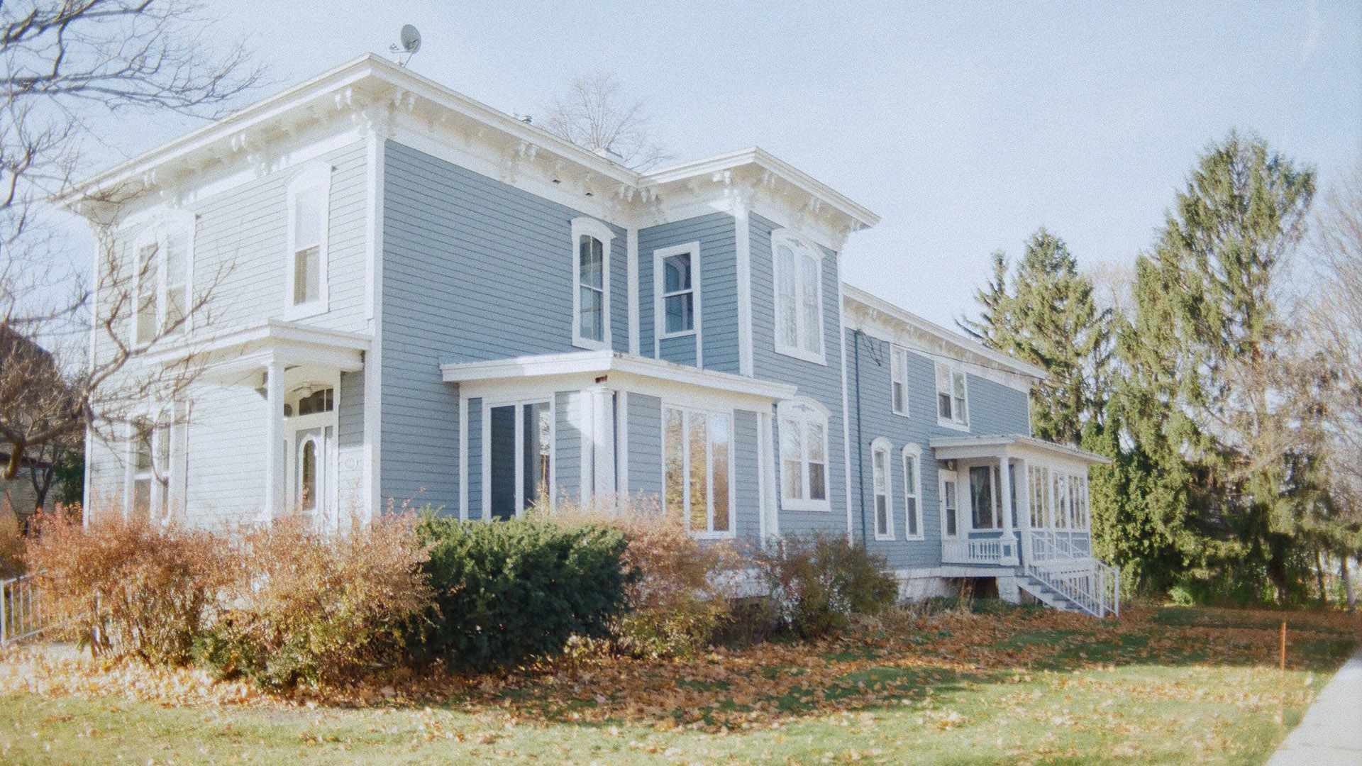 Sun shines on a two-story Italianate house with a satellite dish on its roof, landscape plants along two walls, with fallen leaves on the surrounding lawn and trees in the background.