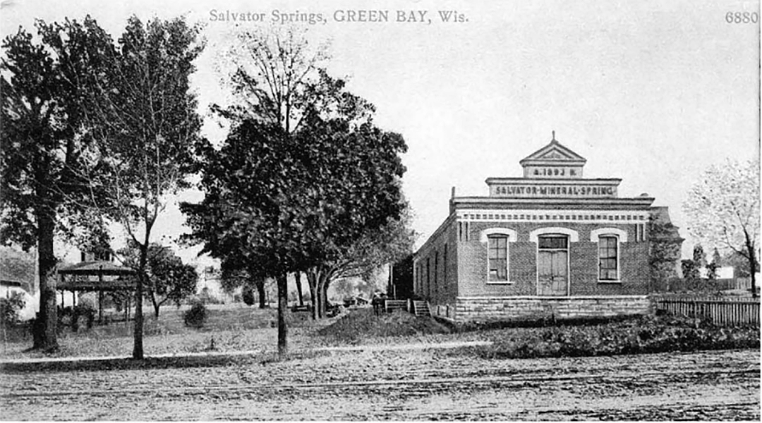 A grayscale postcard shows a stone masonry building with a sign at its top with the year "1892" and the words "Salvator Mineral Springs," with a gazebo and multiple trees to the side, and with a dirt road in the foreground.