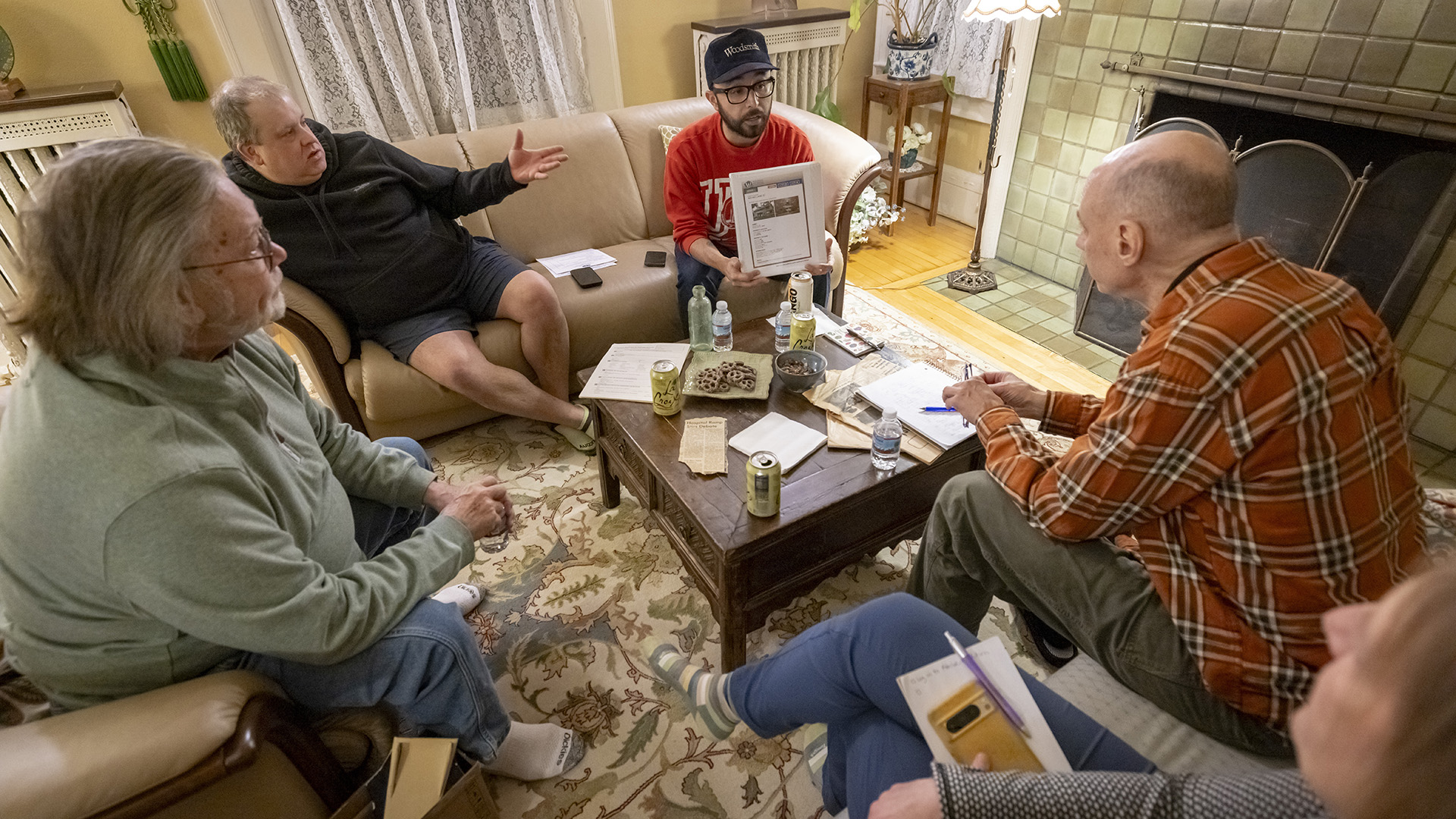 Jim Gucwa, Paul Jacobson, Al Valentin, Skip Heverly and Morgan Fisher sit in multiple couches and chairs around a square table with multiple beverage containers, papers and a plate of cookies on its surface, in a room with a tile-walled fireplace, lamp and radiators on either side of a window with lace curtains.