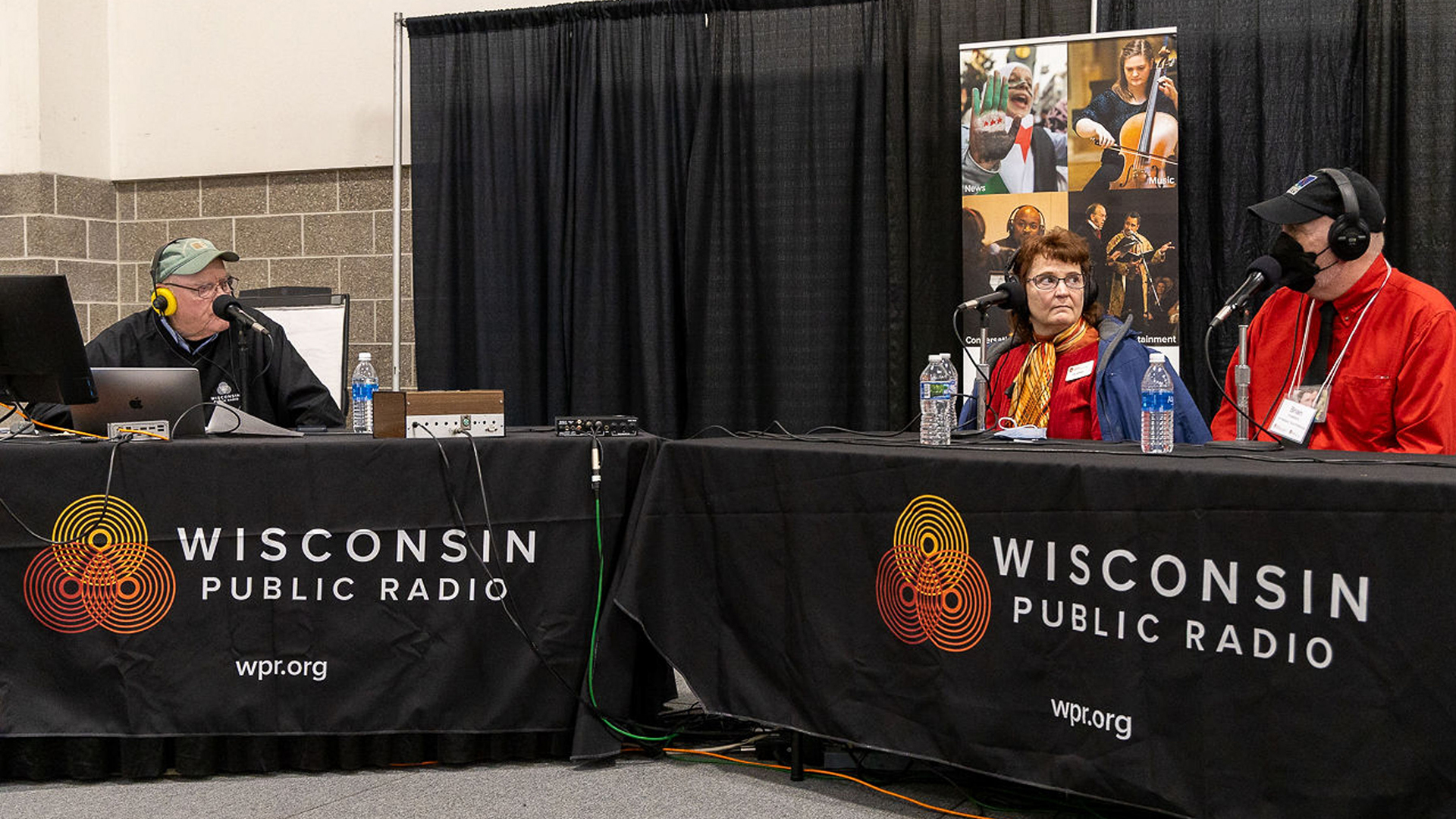 Beth Johnson (left) and Brian Hudelson (right) seated at a table on the right-hand, each with a microphone, engaged in conversation or discussion with an older person on the left-hand side.