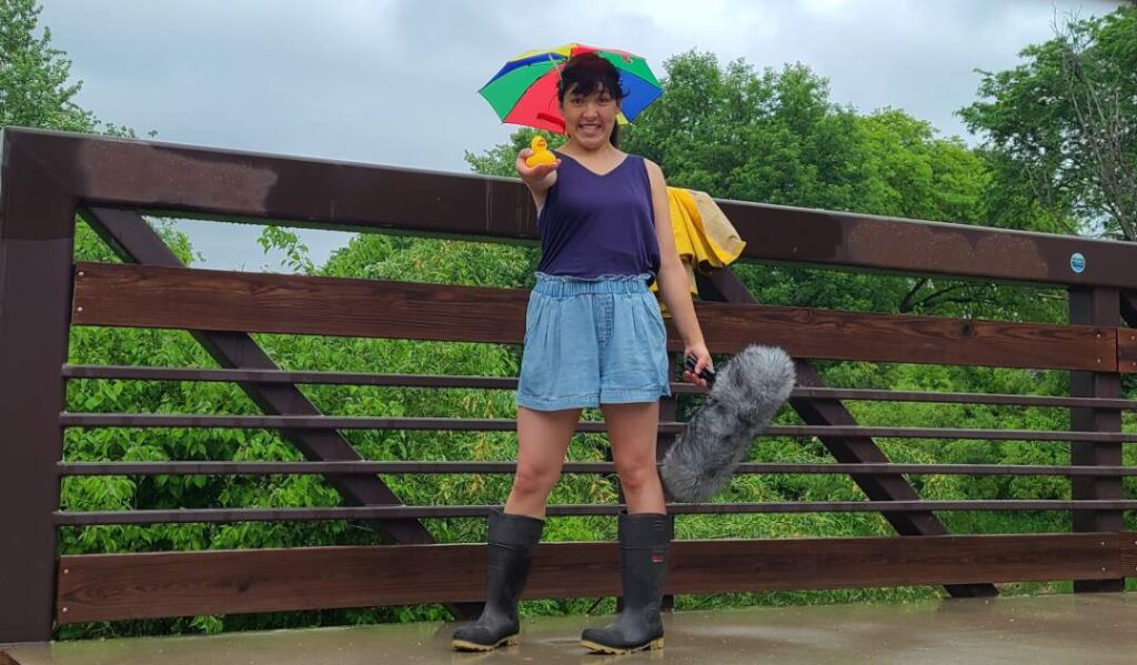 A young woman stands on a bridge with an umbrella hat on in the rain.