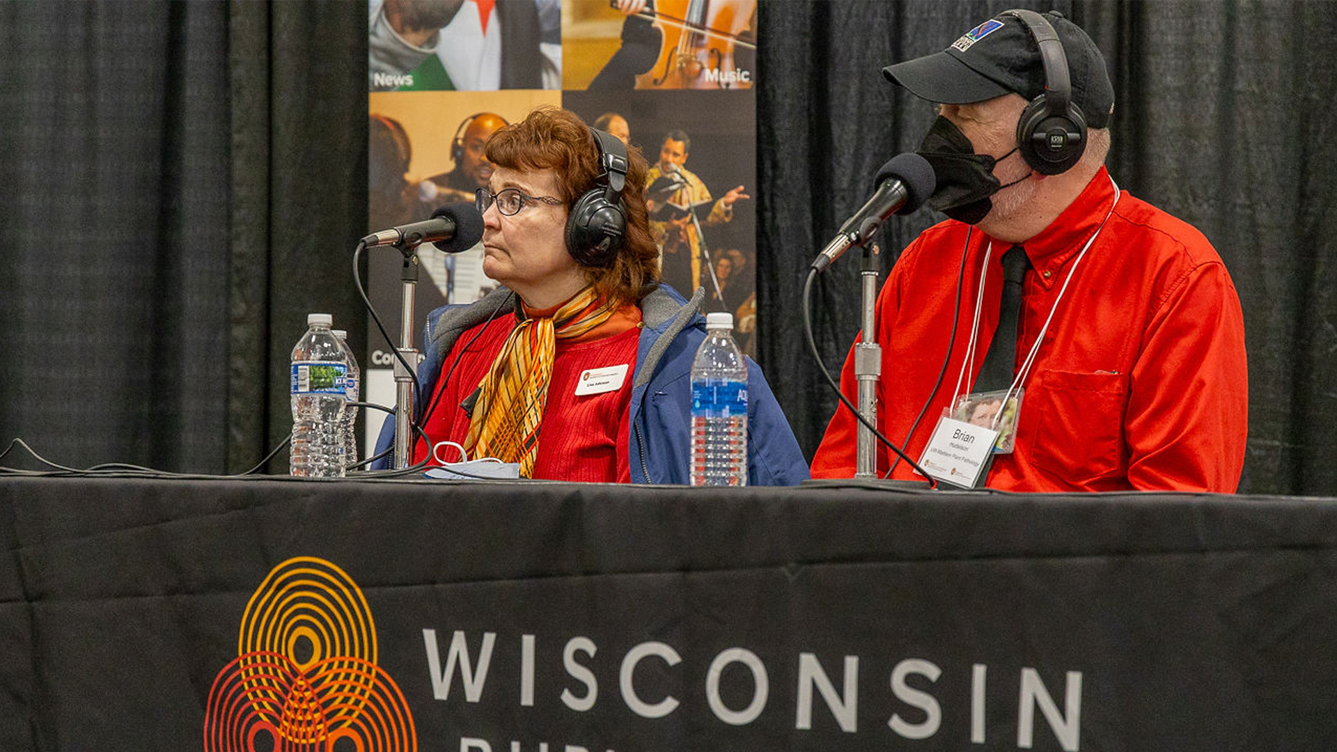 Beth Johnson (left) and Brian Hudelson (right) wearing headphones and speaking into microphones, sit at a table with a "Wisconsin Public Radio" banner. Bottled water is placed on the table.