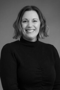 Lindsey Meekhof Smiling woman with shoulder-length wavy hair wearing a dark turtleneck sweater, posed against a plain background in a black-and-white portrait.