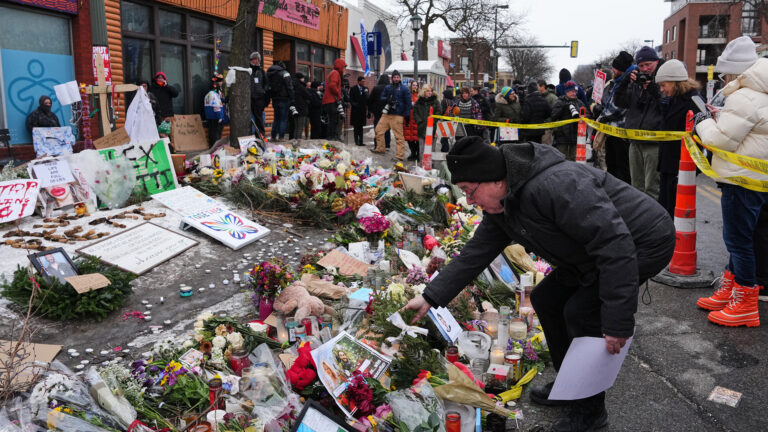 A man wearing a parka and holding a sheet of paper in his left hand places a pine spray with his right hand on a ring-shaped makeshift memorial consisting of flower bouquets, wreaths, votive candles, sign and photos, and other items, with other people standing in the background behind a line of barrier tape strung around multiple traffic cones, with buildings, leafless trees and traffic signals in the background. A man wearing a parka and holding a sheet of paper in his left hand places a pine spray with his right hand on a ring-shaped makeshift memorial consisting of flower bouquets, wreaths, votive candles, sign and photos, and other items, with other people standing in the background behind a line of barrier tape strung around multiple traffic cones, with buildings, leafless trees and traffic signals in the background.