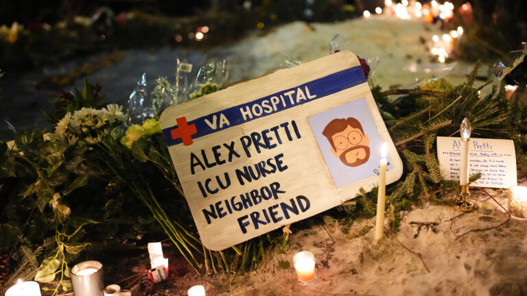A hand-painted sign in the shape of an ID card with a illustration of a portrait of Alex Pretti and the Red Cross symbol next to the words VA Hospital and above the words Alex Pretti, ICU Nurse, Neighbor and Friend rests on top of multiple bouquets of flowers and other foliage, and among multiple burning candles and illuminated electric candles on top of a snowbank, with out-of-focus flowers and candles in the background. A hand-painted sign in the shape of an ID card with a illustration of a portrait of Alex Pretti and the Red Cross symbol next to the words VA Hospital and above the words Alex Pretti, ICU Nurse, Neighbor and Friend rests on top of multiple bouquets of flowers and other foliage, and among multiple burning candles and illuminated electric candles on top of a snowbank, with out-of-focus flowers and candles in the background.