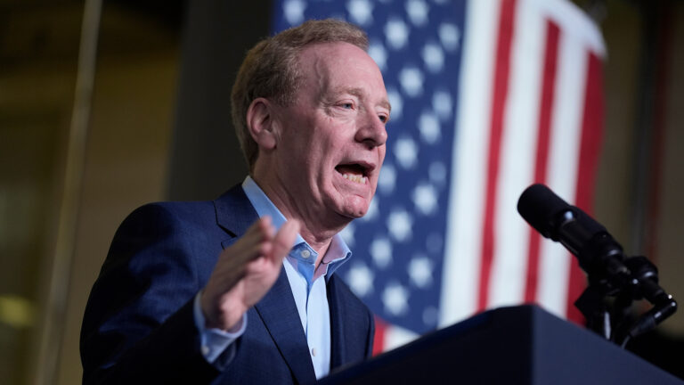 Brad Smith gestures with his right hand, which is out of focus, while speaking into a microphone mounted to the top of a podium, with a U.S. flag in the background.
