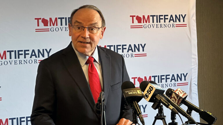 Tom Tiffany speaks while facing multiple mounted microphones with the flags of different media organizations and standing in front of a vinyl backdrop banner showing his campaign logo.