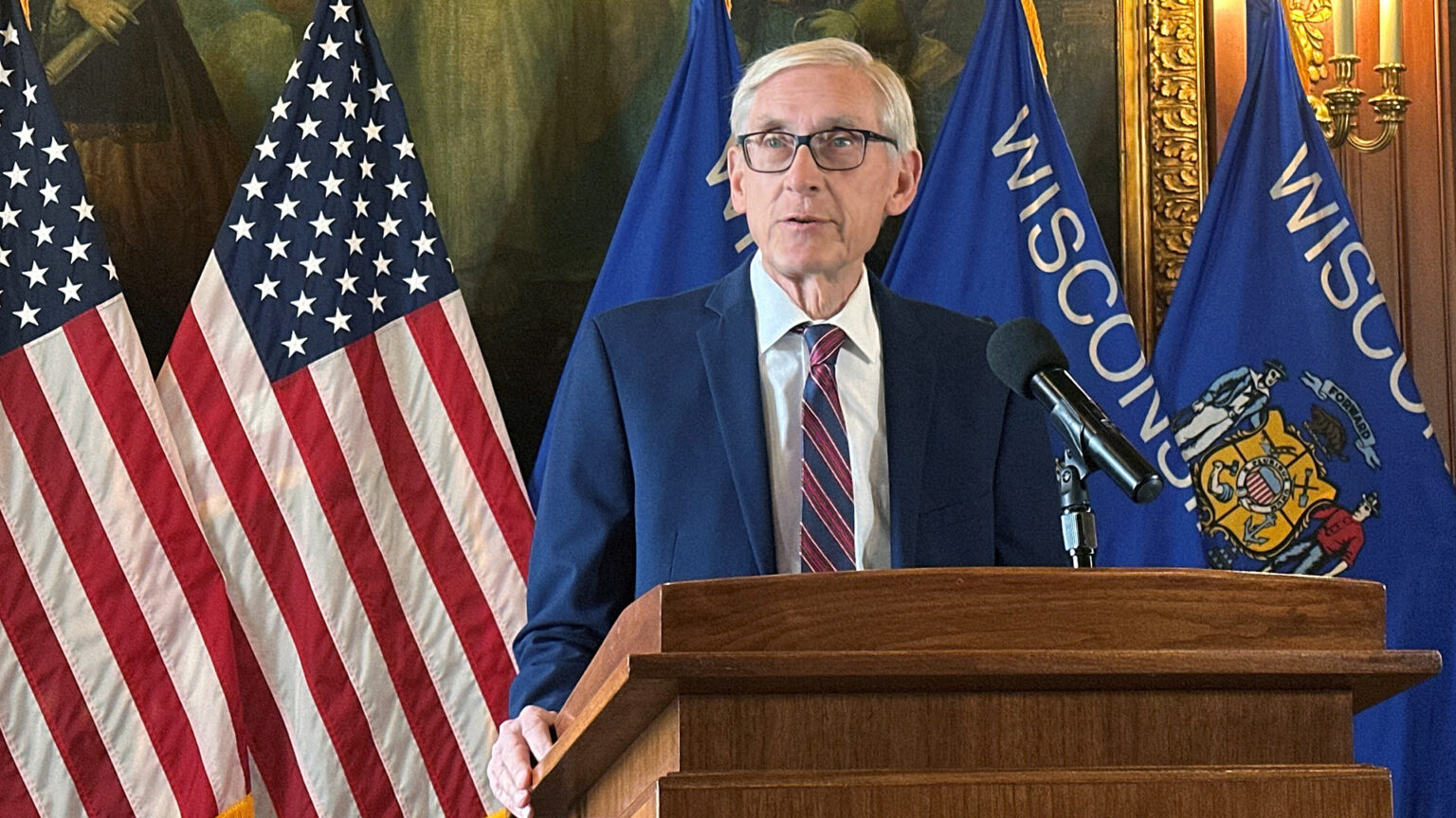 Tony Evers speaks into a microphone mounted to the top of a wood podium while standing in front of a row of U.S. and Wisconsin flags in a room with a large painting in a gilt frame next to a wood-paneled wall.