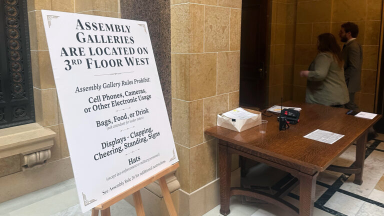 A printed posterboard sign with the title Assembly Galleries Are Located on 3rd Floor West and the text Assembly Rules Prohibit: Cell Phones, Cameras, or Other Electronic Usage is displayed on a wood easel and next to a wood table with stacks of papers and a two-radio on its surface, with two people in the background walking toward a door in a hallway with masonry flooring and walls. A printed posterboard sign with the title Assembly Galleries Are Located on 3rd Floor West and the text Assembly Rules Prohibit: Cell Phones, Cameras, or Other Electronic Usage is displayed on a wood easel and next to a wood table with stacks of papers and a two-radio on its surface, with two people in the background walking toward a door in a hallway with masonry flooring and walls.