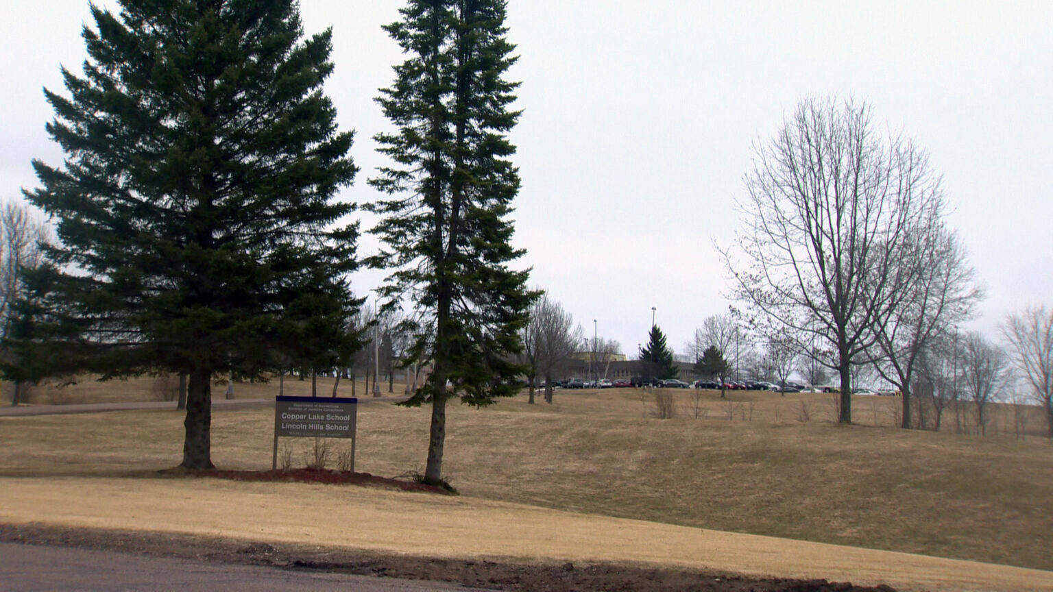 An entrance sign with the words Copper Lake School and Lincoln Hills School stands between two trees and in front of a a series of rolling hills with a tree-lined driveway leading to a parking lot filled with vehicles and multiple buildings surrounded by light poles. An entrance sign with the words Copper Lake School and Lincoln Hills School stands between two trees and in front of a a series of rolling hills with a tree-lined driveway leading to a parking lot filled with vehicles and multiple buildings surrounded by light poles.