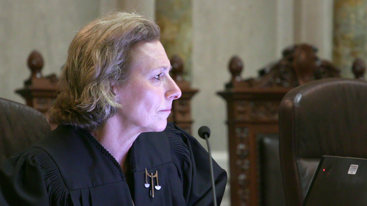 Jill Karofsky sits in a high-backed leather chair and faces a mounted microphone and a monitor with an illuminated power light, with a row of empty high-backed wood and leather chairs in a room with marble pillars and masonry.