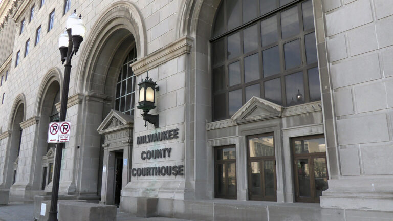 A letter sign reading Milwaukee County Courthouse is affixed to the façade of a building with granite masonry, exterior metal light fixtures, and a door and metal-framed windows set with in arches, with a street light with no parking signs on a sidewalk in front of the entrance. A letter sign reading Milwaukee County Courthouse is affixed to the façade of a building with granite masonry, exterior metal light fixtures, and a door and metal-framed windows set with in arches, with a street light with no parking signs on a sidewalk in front of the entrance.