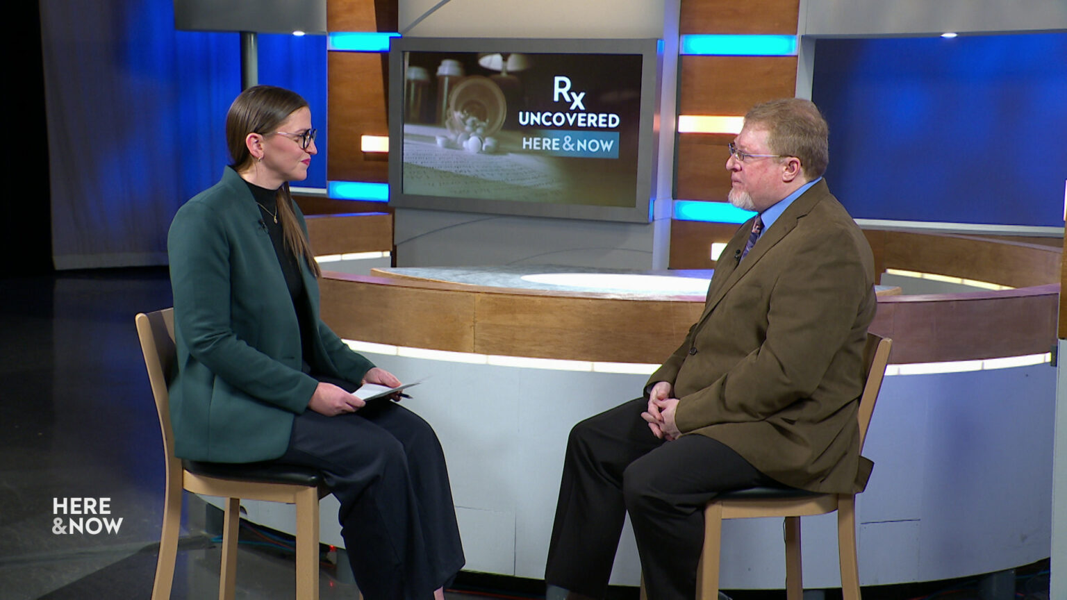 Marisa Wojcik and Rob Gundermann sit on wood chairs in front of the Here & Now set with a monitor showing a graphic with the words Rx Uncovered and Here & Now.