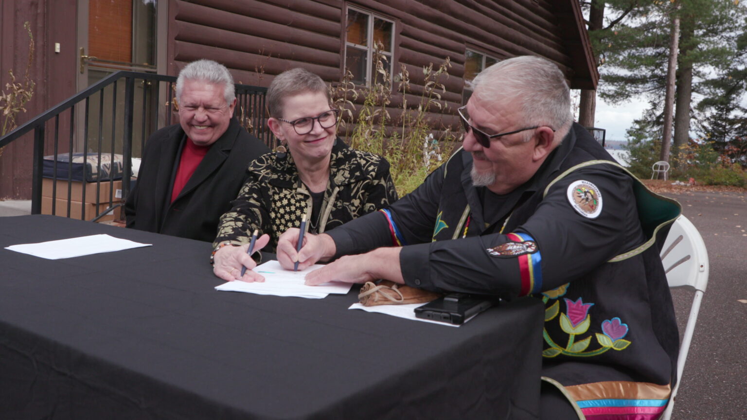John Johnson sits on folding chair at a folding table covered with a tablecloth set up on an asphalt driveway and signs a sheet of paper while seated next to Sue Ernster, who is seated next to Larry Turner in front of a building with log siding, a door and two windows, with trees and a body of water in the background.