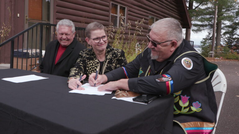 John Johnson sits on folding chair at a folding table covered with a tablecloth set up on an asphalt driveway and signs a sheet of paper while seated next to Sue Ernster, who is seated next to Larry Turner in front of a building with log siding, a door and two windows, with trees and a body of water in the background.