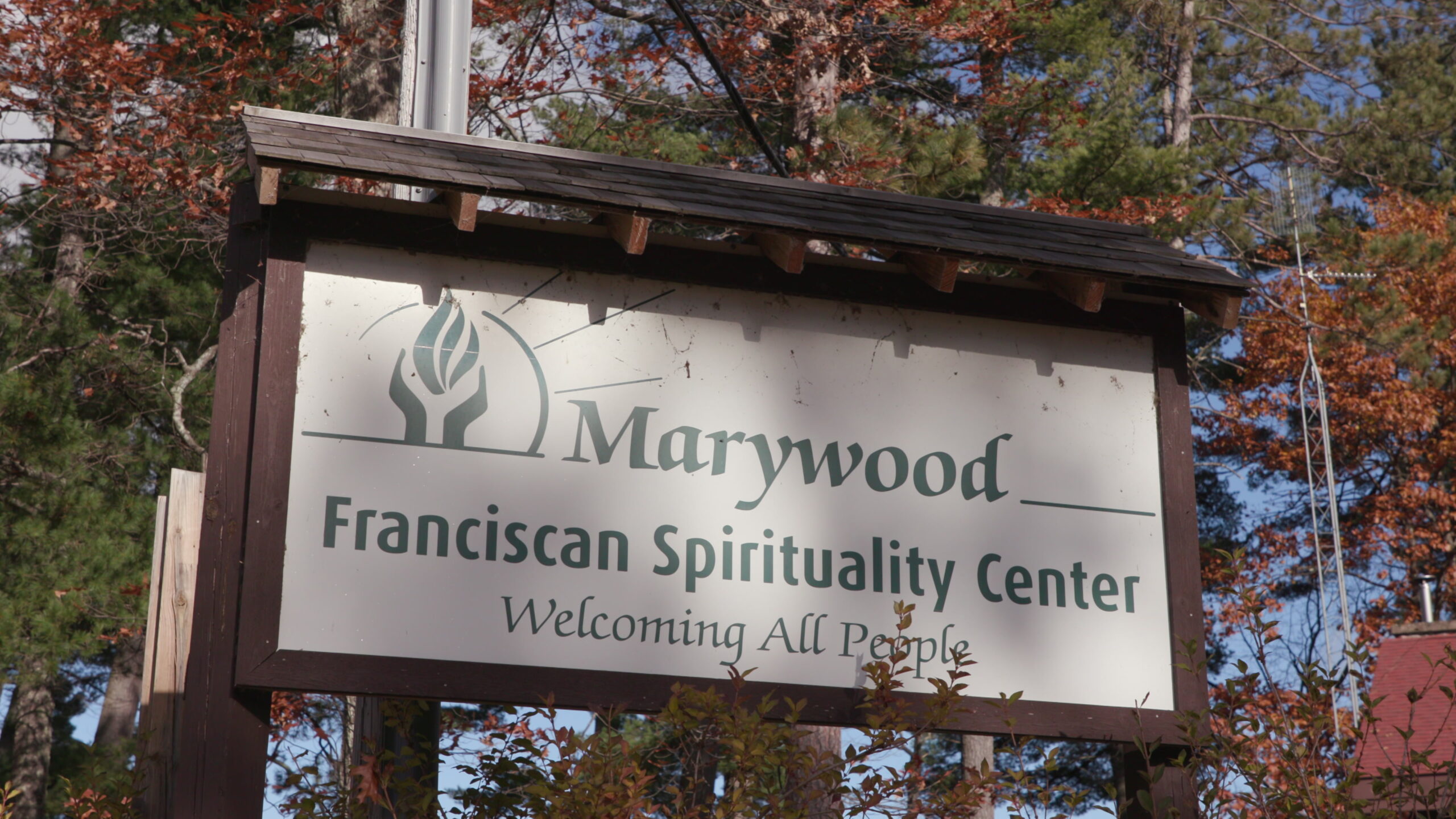 A sign with logo with a fire motif and the words "Marywood Franciscan Spirituality Center" and "Welcoming All People" is attached to the side of a wood stand with a sloped shingle roof, with coniferous trees and deciduous trees with dying leaves in the background.