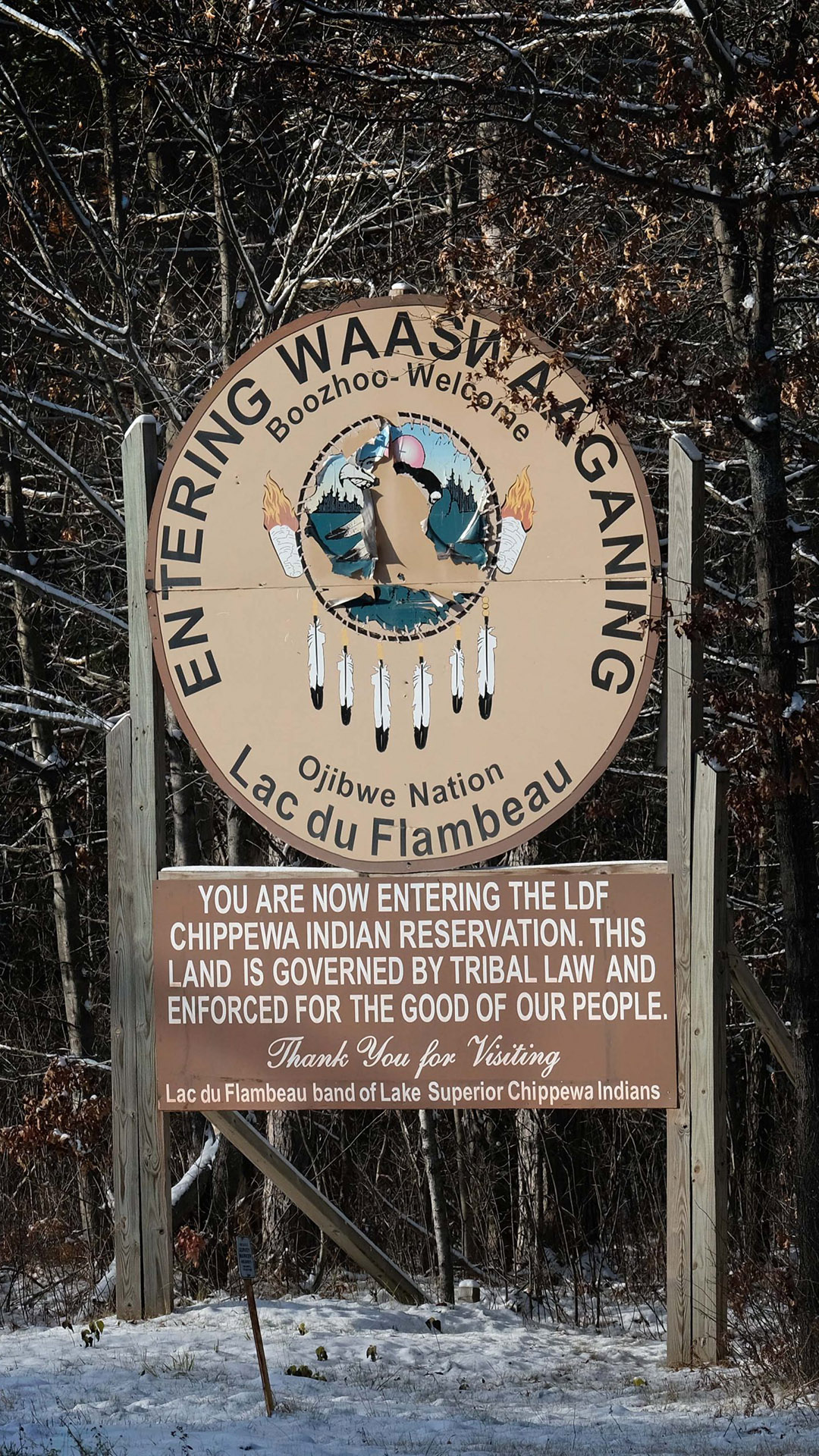 A wood sign with the seal of the Lac du Flambea Band of Lake Superior Chippewa and the words "Entering Waaswaaganing," "Ojibwe Nation Lac du Flambeau," "You are now entering the LDF Chippewa Indian Reservation. This Land is Governed by Tribal Law and Enforced by the Good of Our People," and "Thank you for Visiting" stands in snow front of a wooded area with leafless trees.