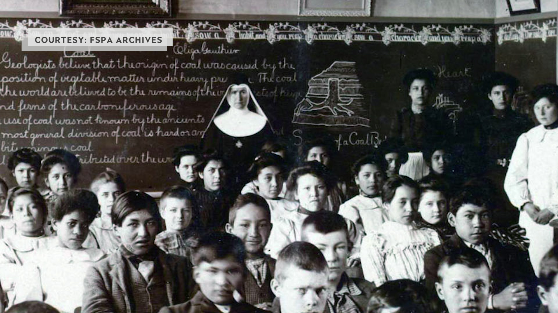A grayscale photo shows seated and standing students and a nun wearing a habit standing in a room with a wall-to-wall chalkboard with lines of cursive writing and a drawing of a tree stump, with a graphic label in the upper-right corner reading "Courtesy: FSPA Archives."