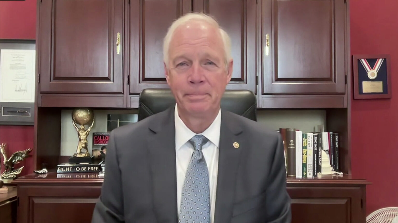 Ron Johnson listens while sitting in front of wood cabinet with multiple books and statues on a shelf in front of a wall with multiple framed items displayed on its surface.