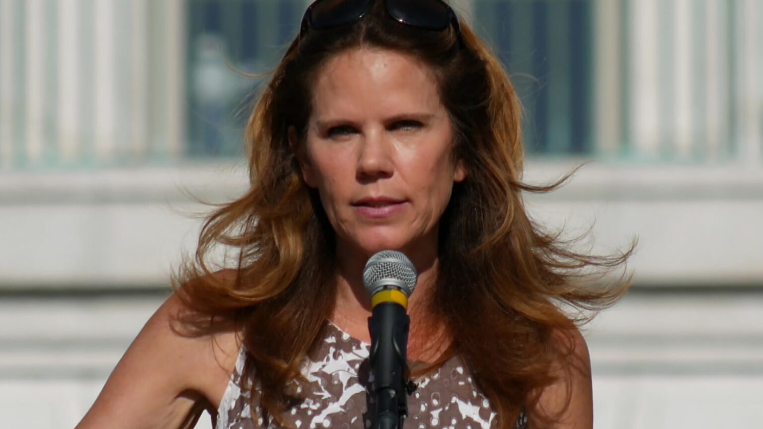 Chris Taylor speaks into a microphone while standing outside, with an out-of-focus window with vertical blinds in the background.