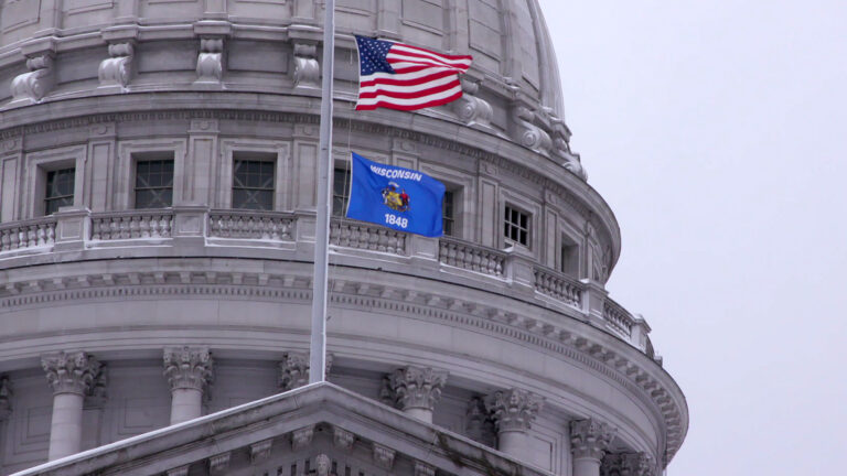 The U.S. and Wisconsin flags fly at half staff on a flagpole mounted to the top of a pediment of a masonry building in front of a dome.
